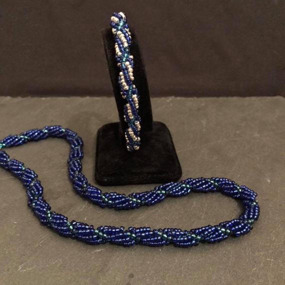 Necklace and bracelet versions of the spiral rope pattern using seed beads. The bracelet is silver and blue and is shown on a black velvet bracelet stand. The necklace is just blue and is lying n front of the bracelet stand on a grey slate.