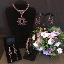 Purple pendant jewellery set. A necklace with a removable pendant, a matching bracelet, and three pairs of matching earrings. Purple and white flowers complete the scene.
