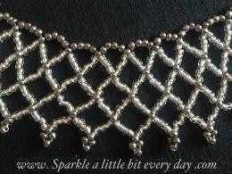 Close up view of the seed bead netted necklace made from silver and grey beads and lying on a grey fabric background.