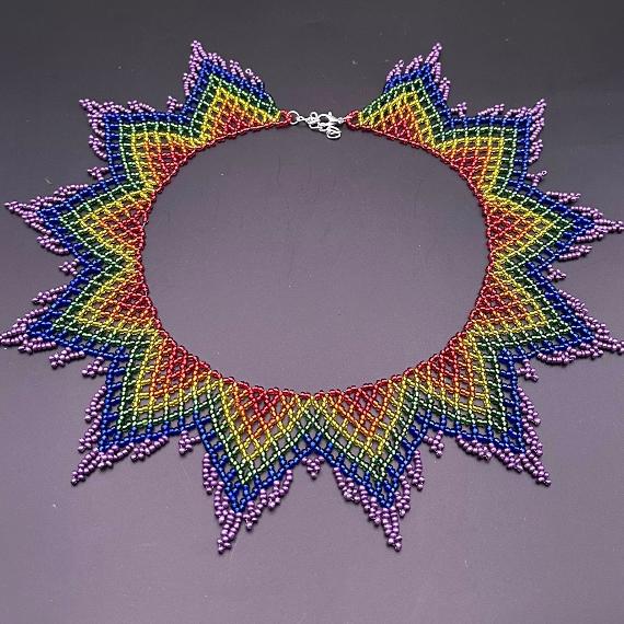 A photo of a rainbow coloured lace collar necklace on a velvet stand with a white lace background.