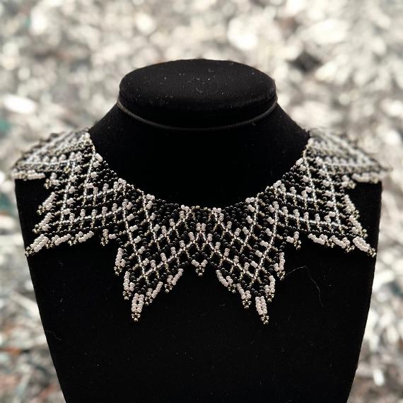 A lace collar necklace made from black, grey, silver, and white beads in front of a silver tinsel background..